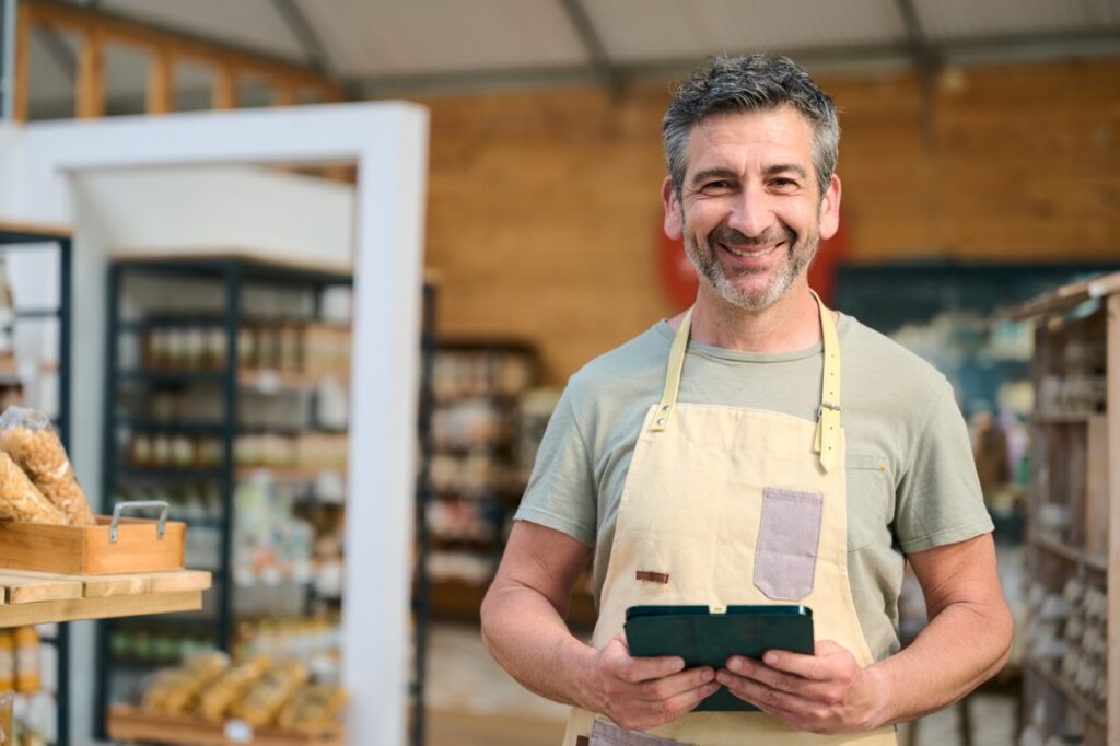 Grocery store owner in an apron reviewing data on a tablet before deciding to open a second grocery store
